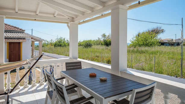 A covered outdoor area with a table and chairs, a hammock, and a view of a house and greenery. - Serenity Apartments (Corfu)
