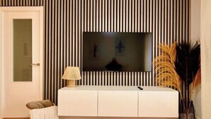 A modern living room with a white entertainment unit, a television mounted on a striped wall, a small side table with a lamp, and decorative plants. - Front line beach - sea view - terrace (Fuengirola)