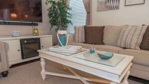 A living room with a glass-top coffee table, a sofa with striped cushions, a television, a plant, and a small cabinet with a dish rack.