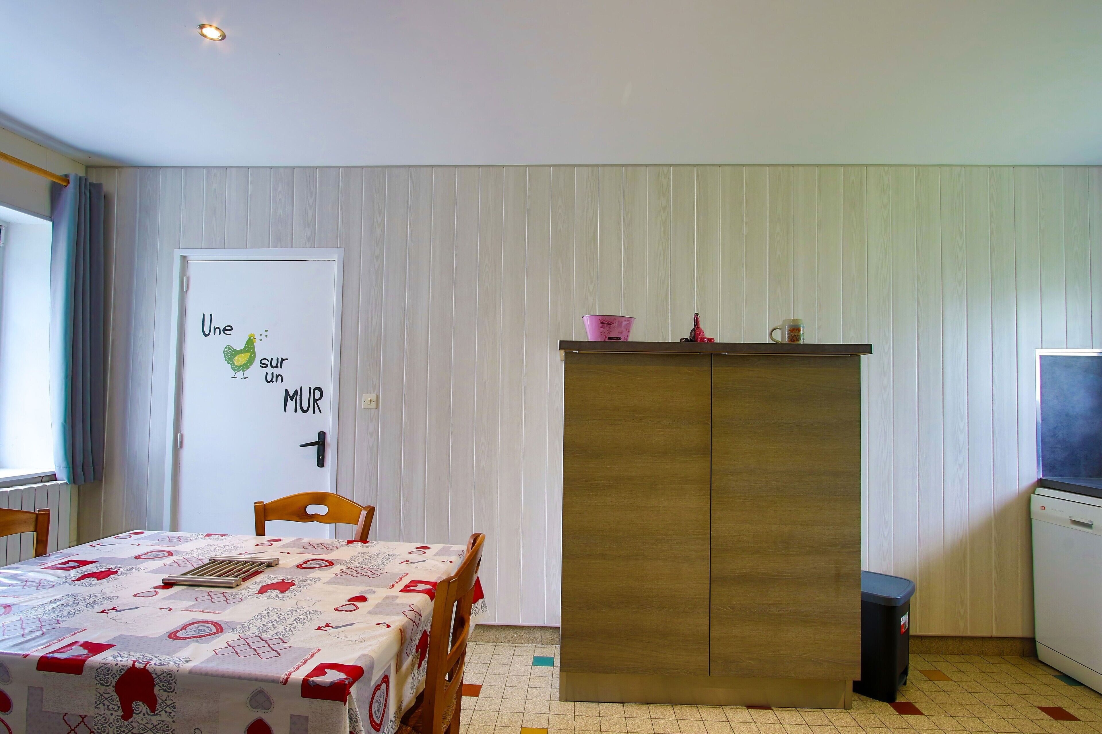 Une pièce comprenant une table à manger, des chaises, une armoire, une poubelle et un mur sur lequel est affiché un panneau.