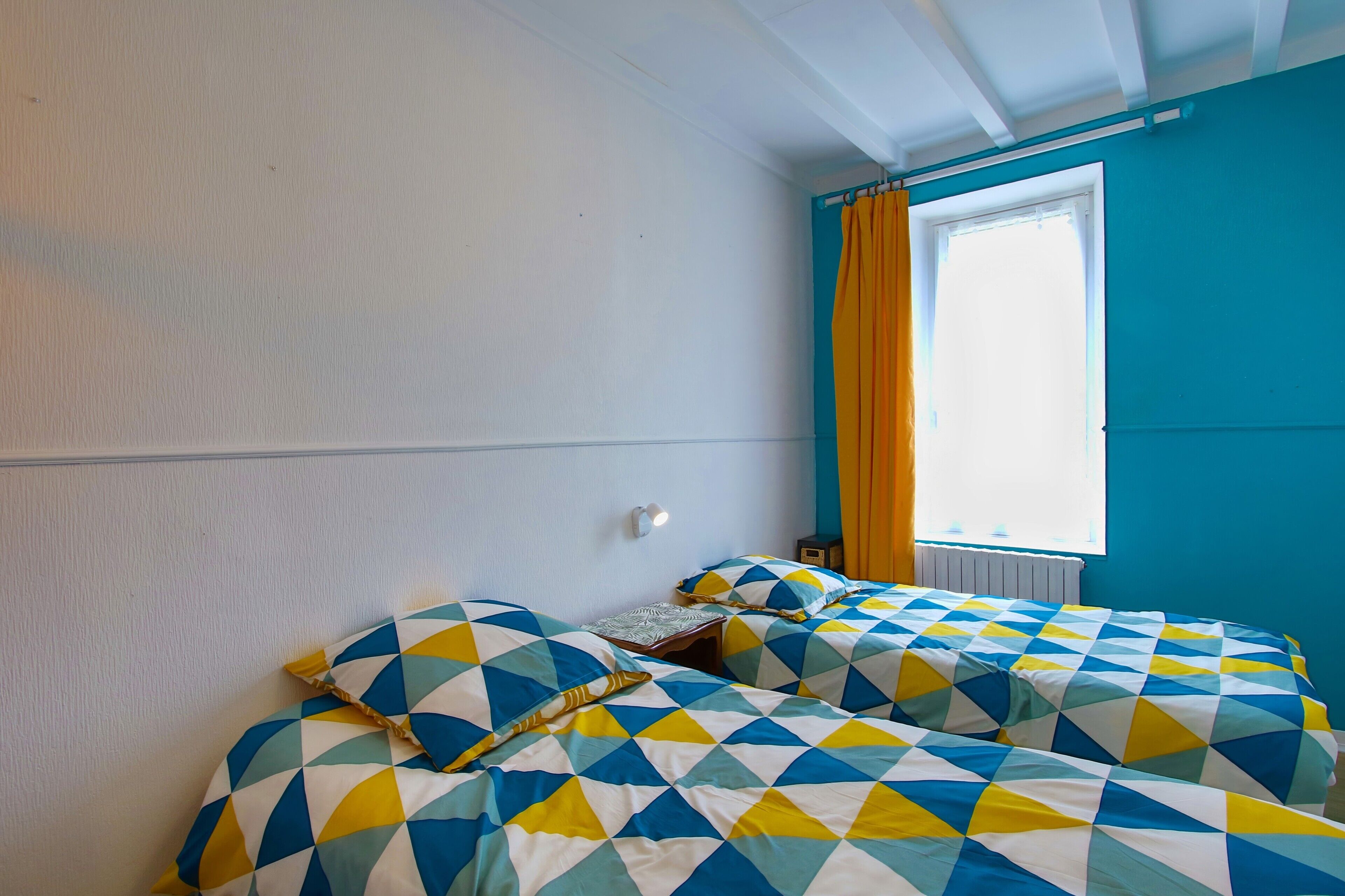 Una habitación con dos camas, una ventana con cortinas, un radiador y una lámpara fijada a la pared.