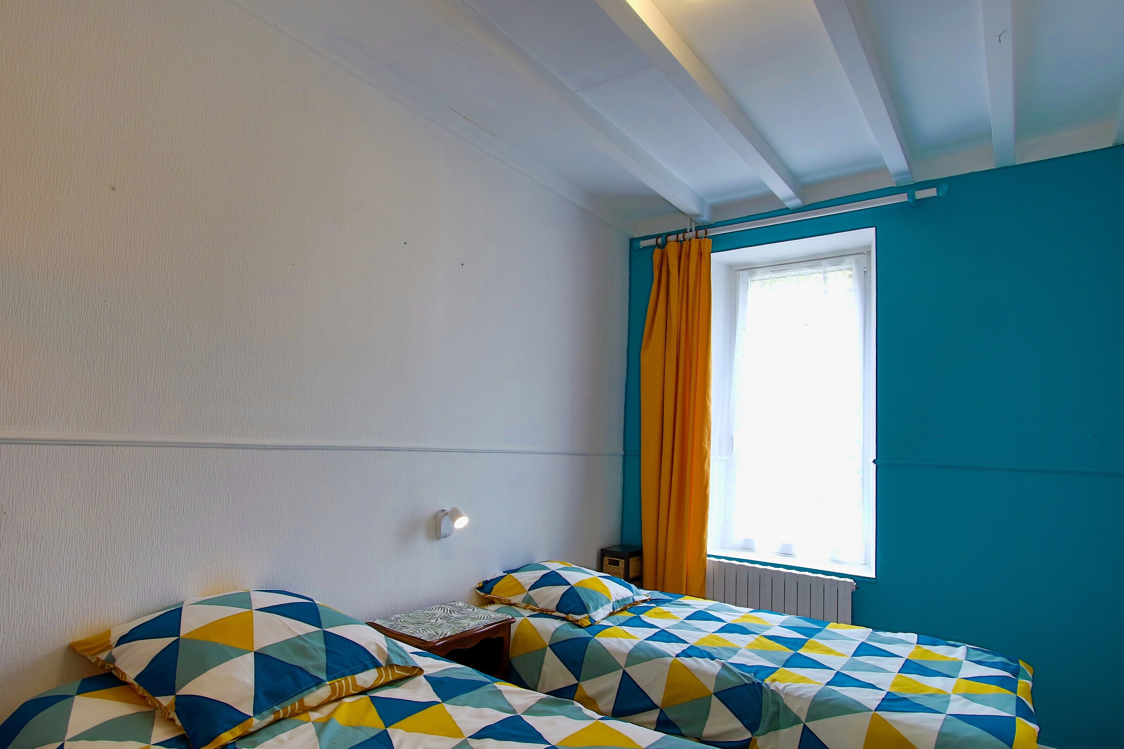 A room with two beds, a window with curtains, a wall-mounted light, and a radiator.