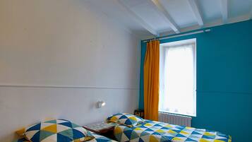 A room with two beds, a window with curtains, a wall-mounted light, and a radiator.