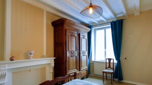 A room with a bed, a wooden wardrobe, a chair, a window with curtains, and a hanging light fixture.