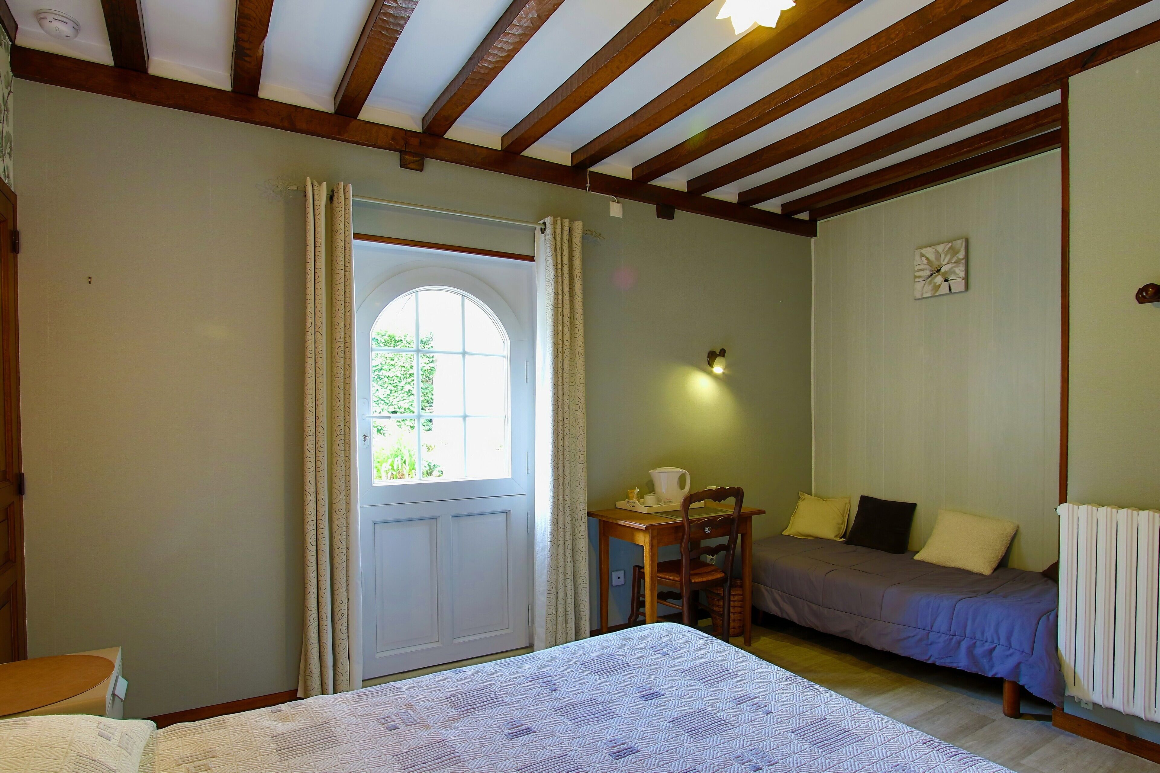 Een kamer met een bed, een tafel met een kopje, een stoel, een radiator en een deur met gordijnen.