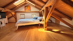 A loft bedroom with a sloped ceiling, wooden beams, a bed, a TV on a stand, and two windows.