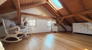 A high-ceilinged attic room with wooden beams, a rocking chair, a bedside table with a lamp, a dresser, and two windows.