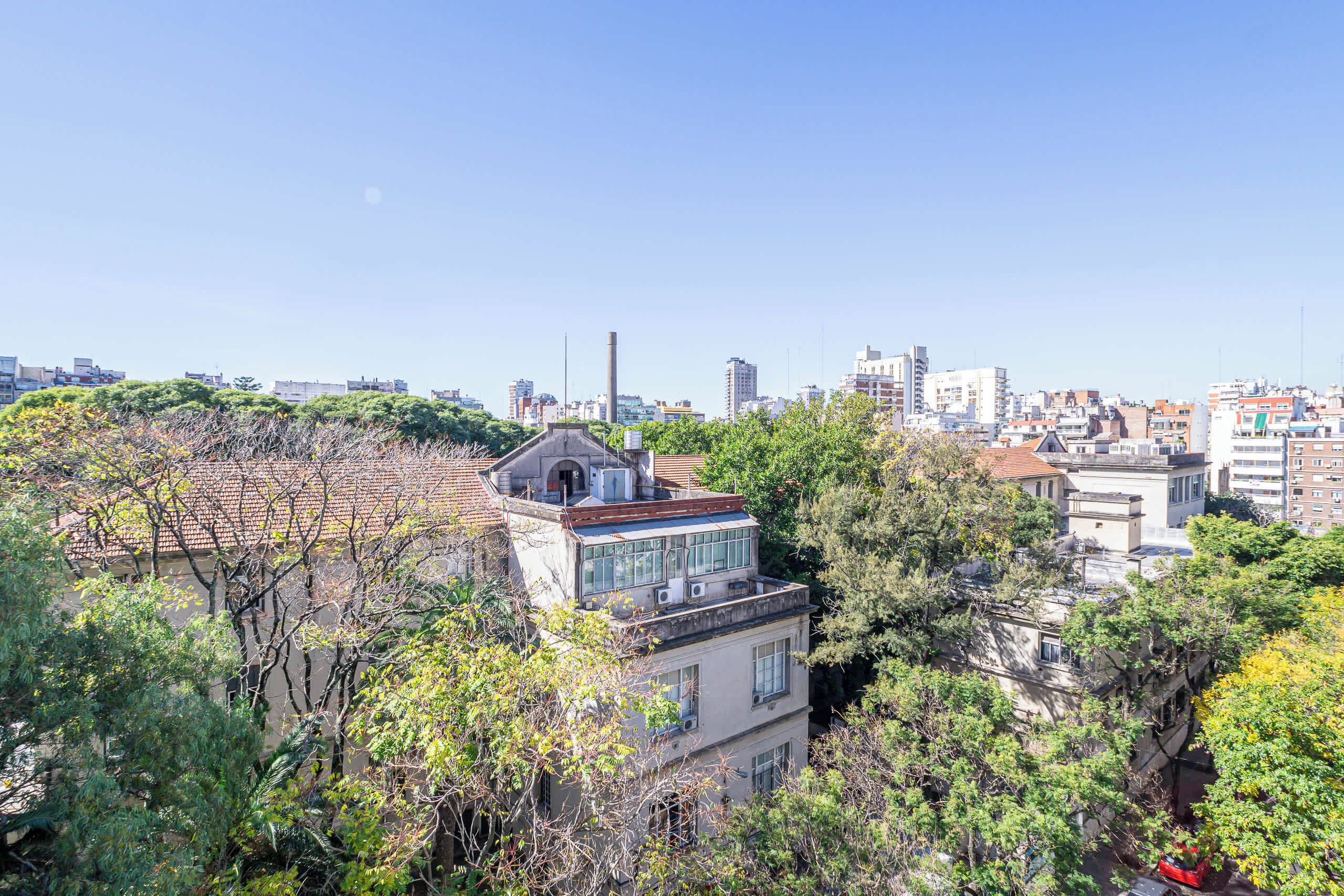 Un edificio de varios pisos con terraza en la azotea, rodeado de árboles y otros edificios.
