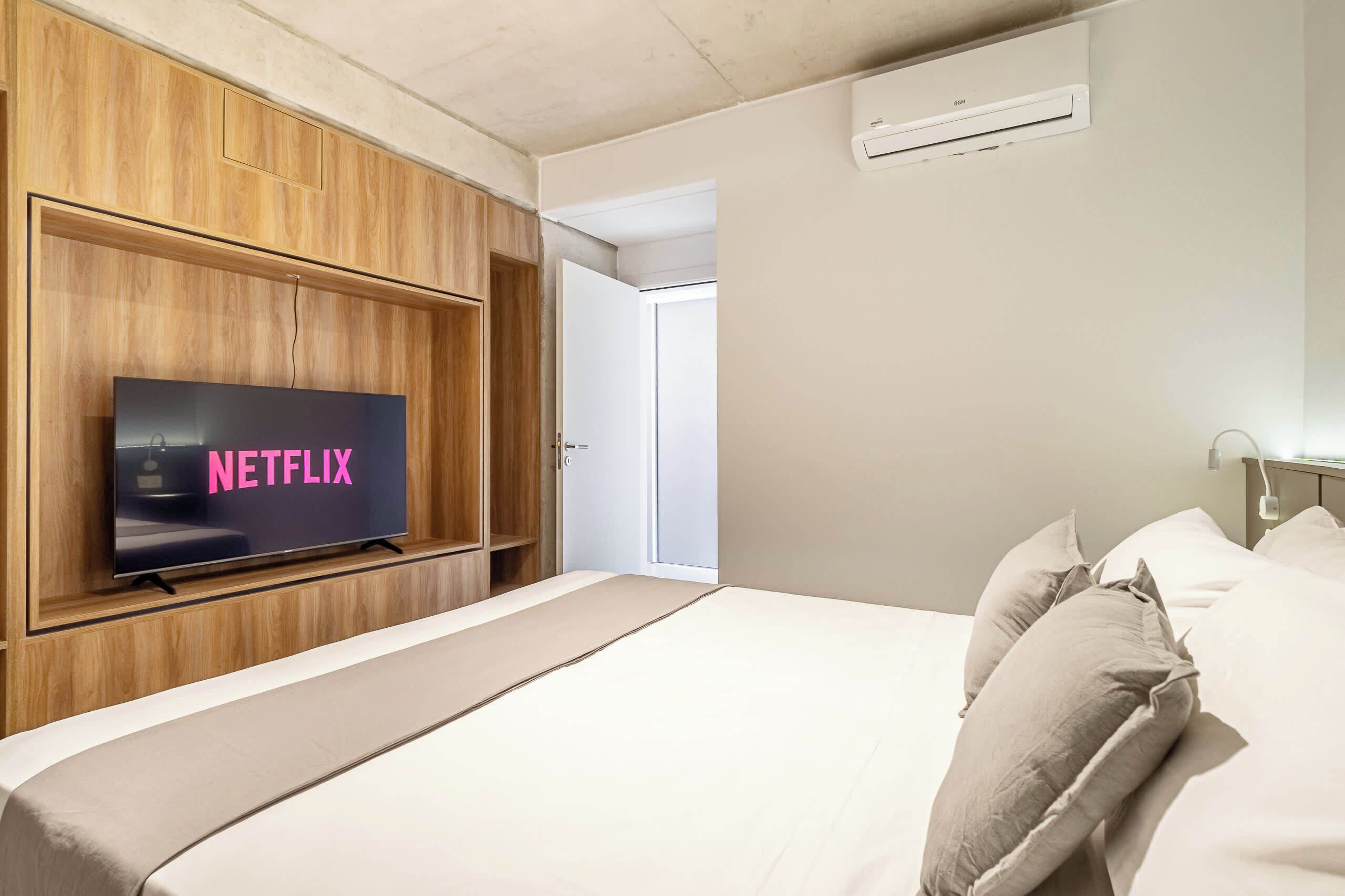 Habitación de hotel con una cama, televisor montado en un panel de madera, una puerta y un equipo de aire acondicionado.