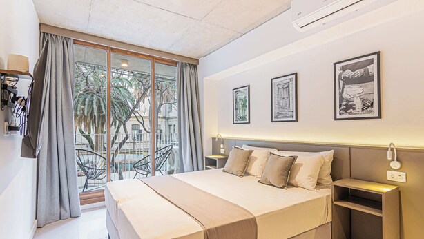 Ein modernes Hotelzimmer mit einem großen Bett, einem Nachttisch, einem kleinen Schreibtisch und einem Balkon mit Blick auf Palmen.