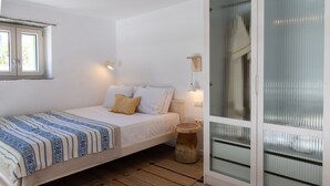 A bedroom with a bed, a window, a bedside table, a lamp, and a door with frosted glass panels. - Veggera Living, Tinos (Pyrgos)