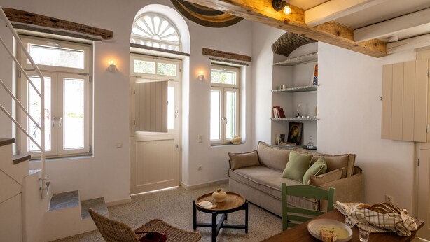 A living room with a sofa, a dining table, a chair, a staircase, and a kitchen area with shelves. - Veggera Living, Tinos (Pyrgos)