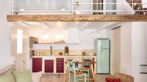 A spacious loft with a high ceiling, exposed beams, a dining area with a table and chairs, a kitchen with cabinets and a refrigerator, and a cozy seating area with a sofa and a chair. - Veggera Living, Tinos (Pyrgos)