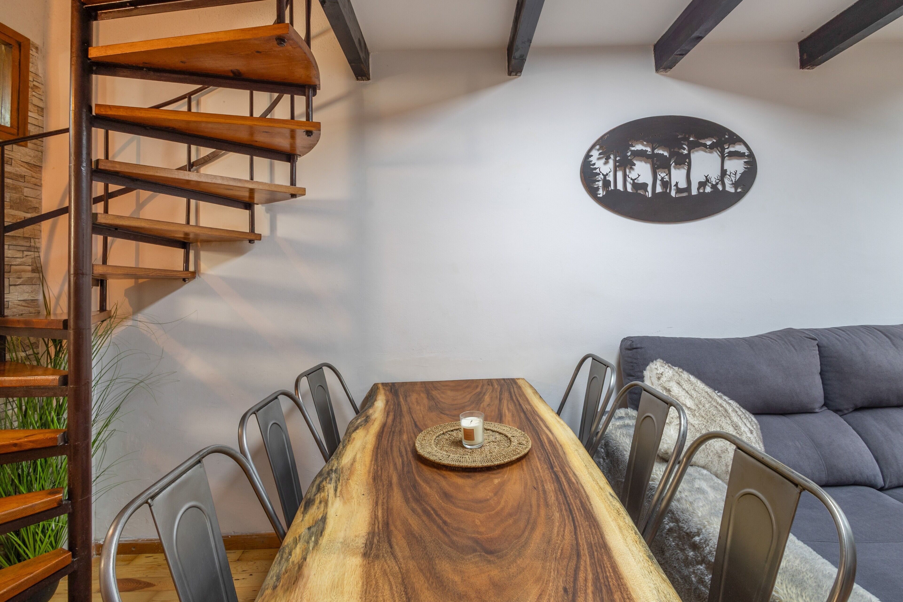A dining area with a wooden table, metal chairs, a glass of water, a woven placemat, a gray sofa, and a wall-mounted artwork.