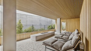 Ein moderner Außenbereich mit einer Sitzecke, einer Couch und Kissen, einem Feuerkorb und Blick auf Grünflächen sowie ein Gebäude im Hintergrund.