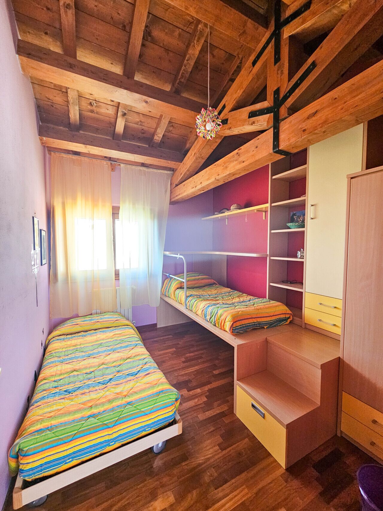 A room with two bunk beds, wooden beams, a hanging light fixture, and a window with sheer curtains.
