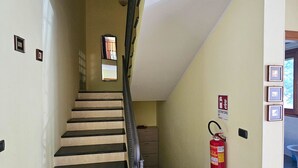 Eine Treppe mit schwarzen Stufen führt zu einer Zwischenebene. Ein Feuerlöscher ist an der Wand befestigt, und eine Tür weist mit einem Schild auf einen Notausgang hin.