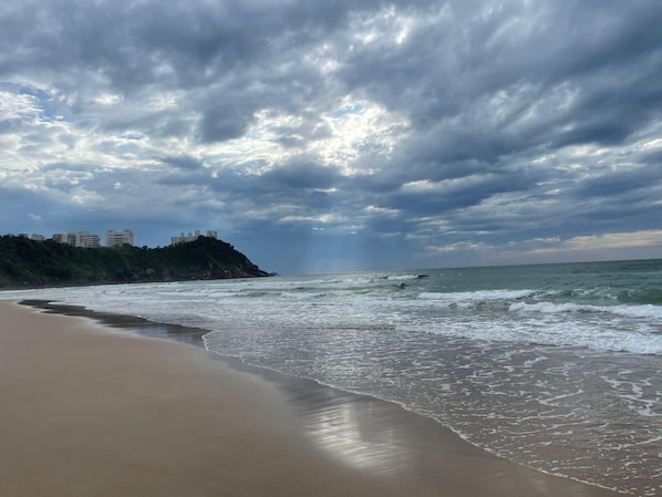 A beach with waves, a sandy shore, and a distant city skyline under a cloudy sky.