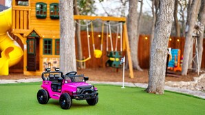 Een speelgoedauto op kunstgras, een houten schommel en een gele glijbaan in een speeltuin.