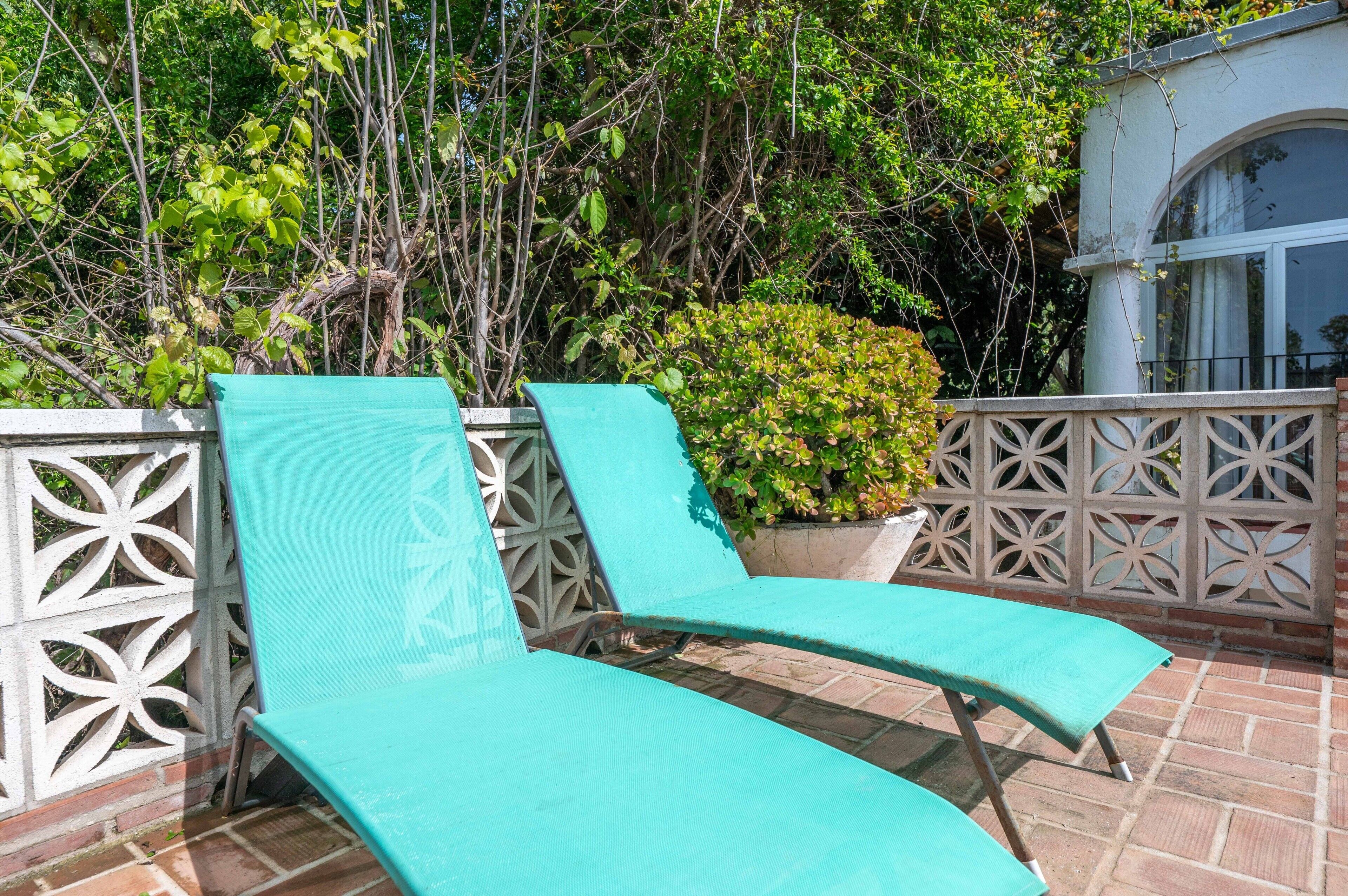 Deux fauteuils de jardin turquoise sont placés sur une terrasse en briques, agrémentée d’une clôture décorative et d’un pot de fleurs.