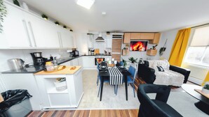 A modern kitchen with white cabinets, a dining area with a striped tablecloth, and a living room with a black sofa and a fireplace. - Stylish 2BR Apartment on Brick Lane – Near Shoreditch (London)