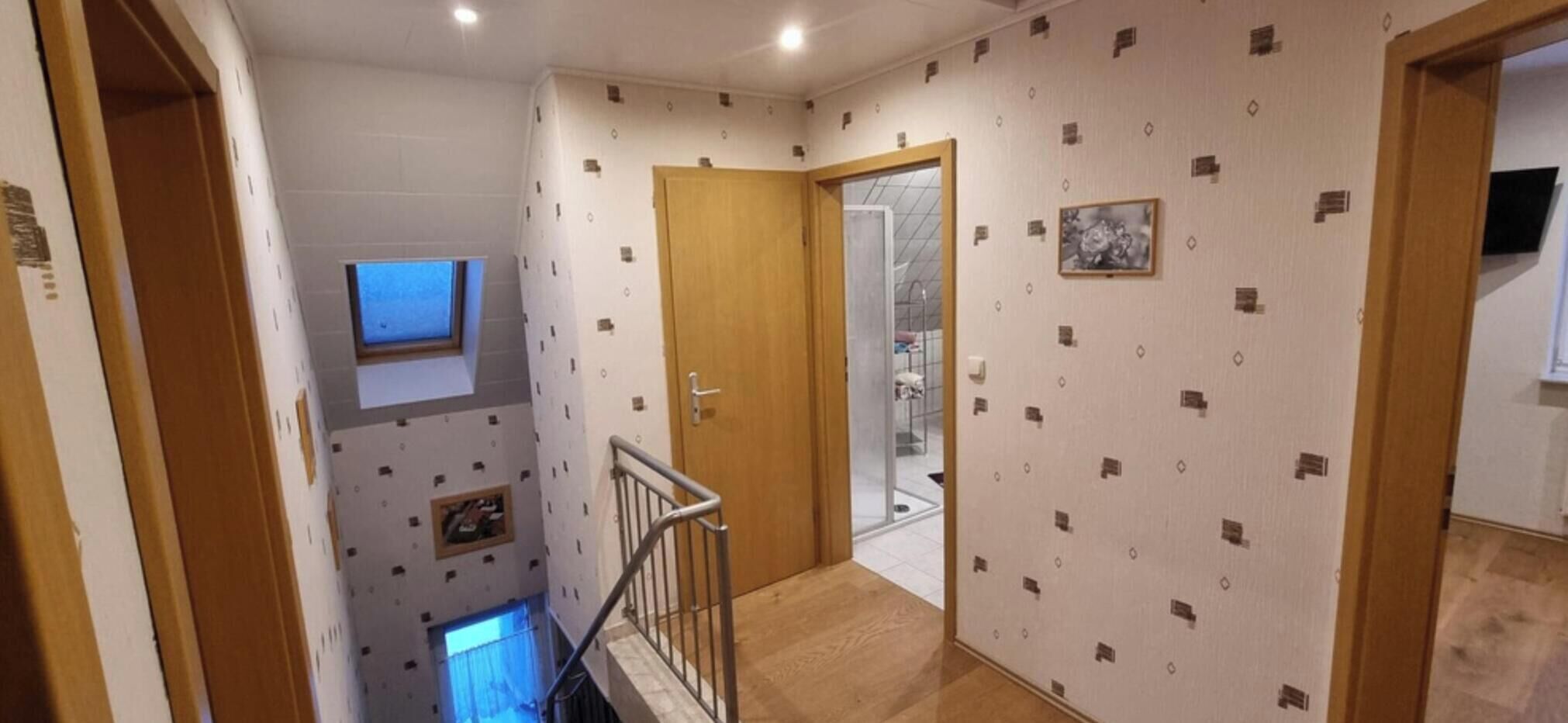 Een trap die leidt naar een kamer met behang met een patroon, een klein raam en een deur naar een andere kamer.