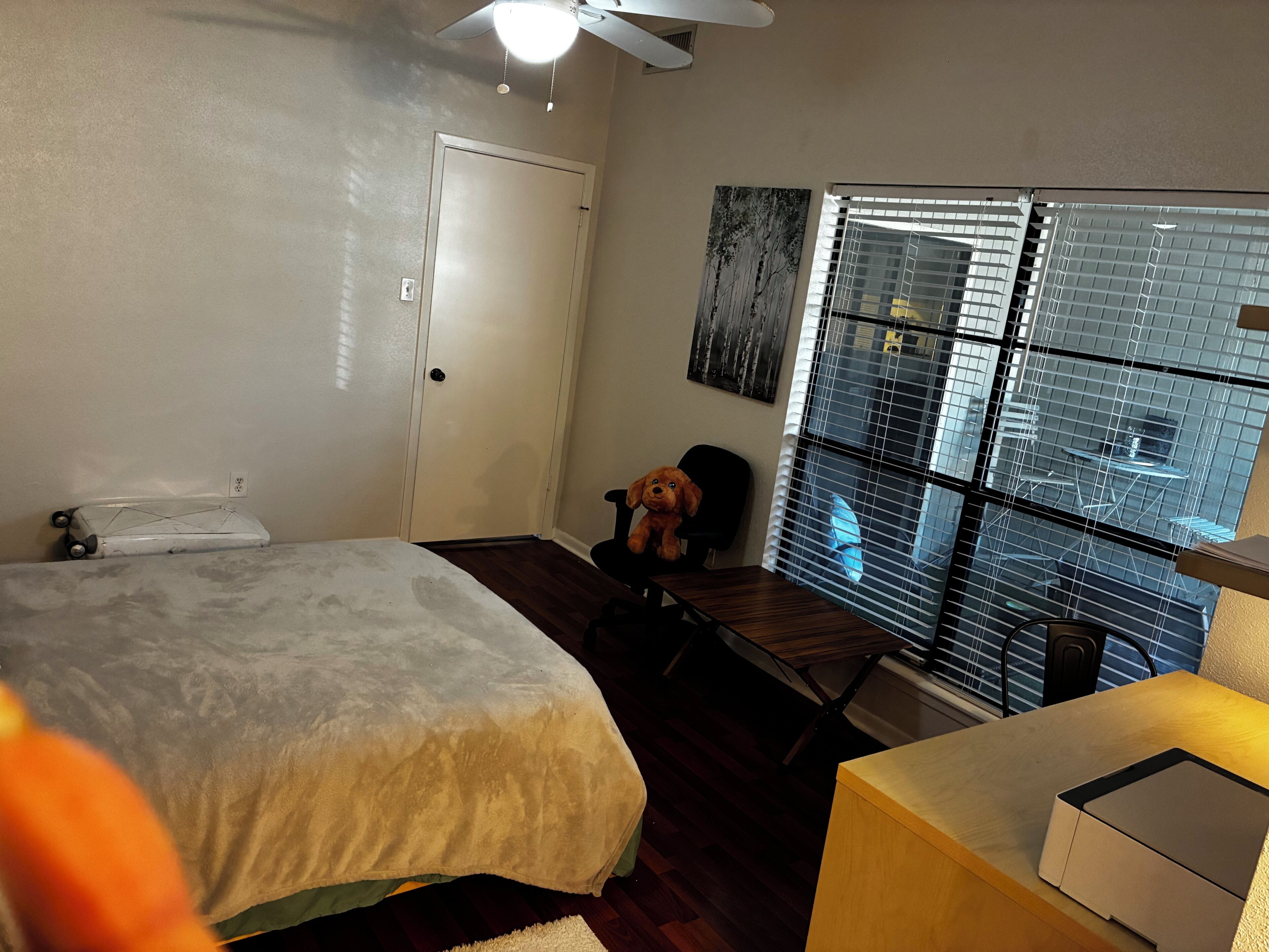 A bedroom with a bed, a desk, a chair, a window with blinds, and a ceiling fan.