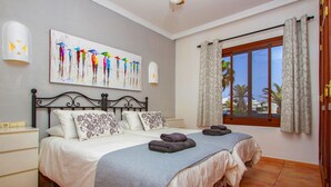 Une chambre à coucher avec un lit, une table de chevet, un tableau, une fenêtre avec des rideaux et une vue sur des palmiers.