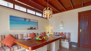 Un coin repas avec une longue table en bois, des sièges rembourrés, un panier de fruits et un tableau représentant une scène de plage.