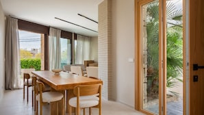 A modern dining area with a wooden table and chairs, large windows, and a view of greenery outside. - Villa Lliris - Four Bedroom Villa, Sleeps 10 (Platja d'Aro)