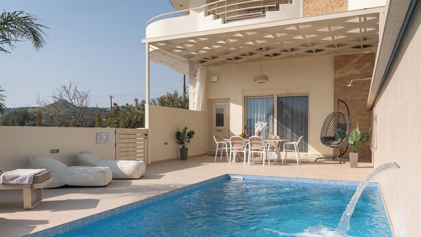 Ein Pool mit Wasserspiel, eine Sitzgruppe mit Kissen, ein Tisch mit Stühlen und ein Hängesessel.