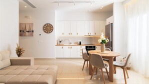 Ein modernes Wohnzimmer mit einem beigen Sofa, einem Essbereich mit rundem Tisch und Stühlen, einer Küche mit weißen Schränken und einer Uhr an der Wand.