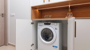 Eine kompakte Waschmaschine in einem kleinen Hauswirtschaftsraum mit einem Holzbodenregal darüber und einem weißen Schrank mit zwei Griffen.