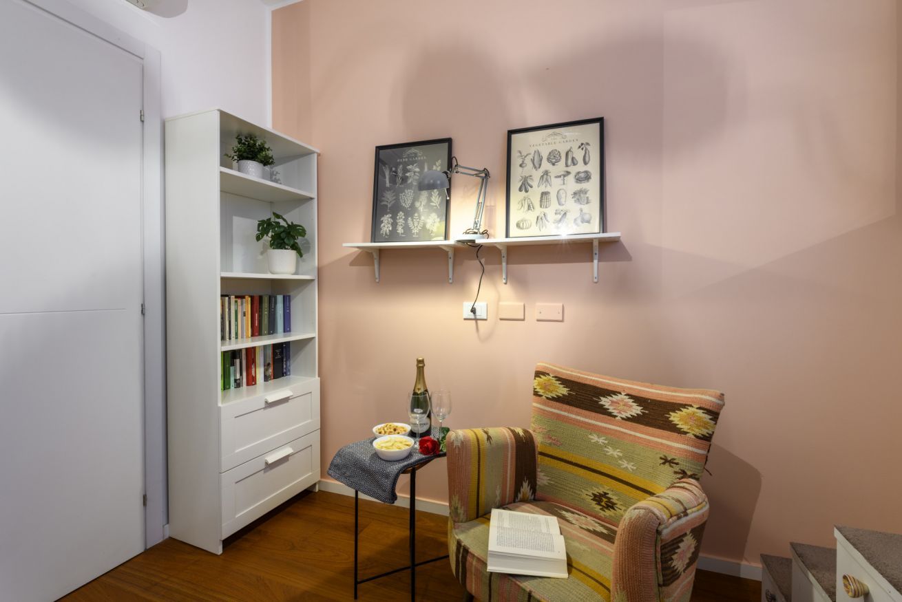 A room with a white bookshelf, a small table with a bottle and glasses, a patterned armchair, and two wall-mounted shelves with framed pictures.