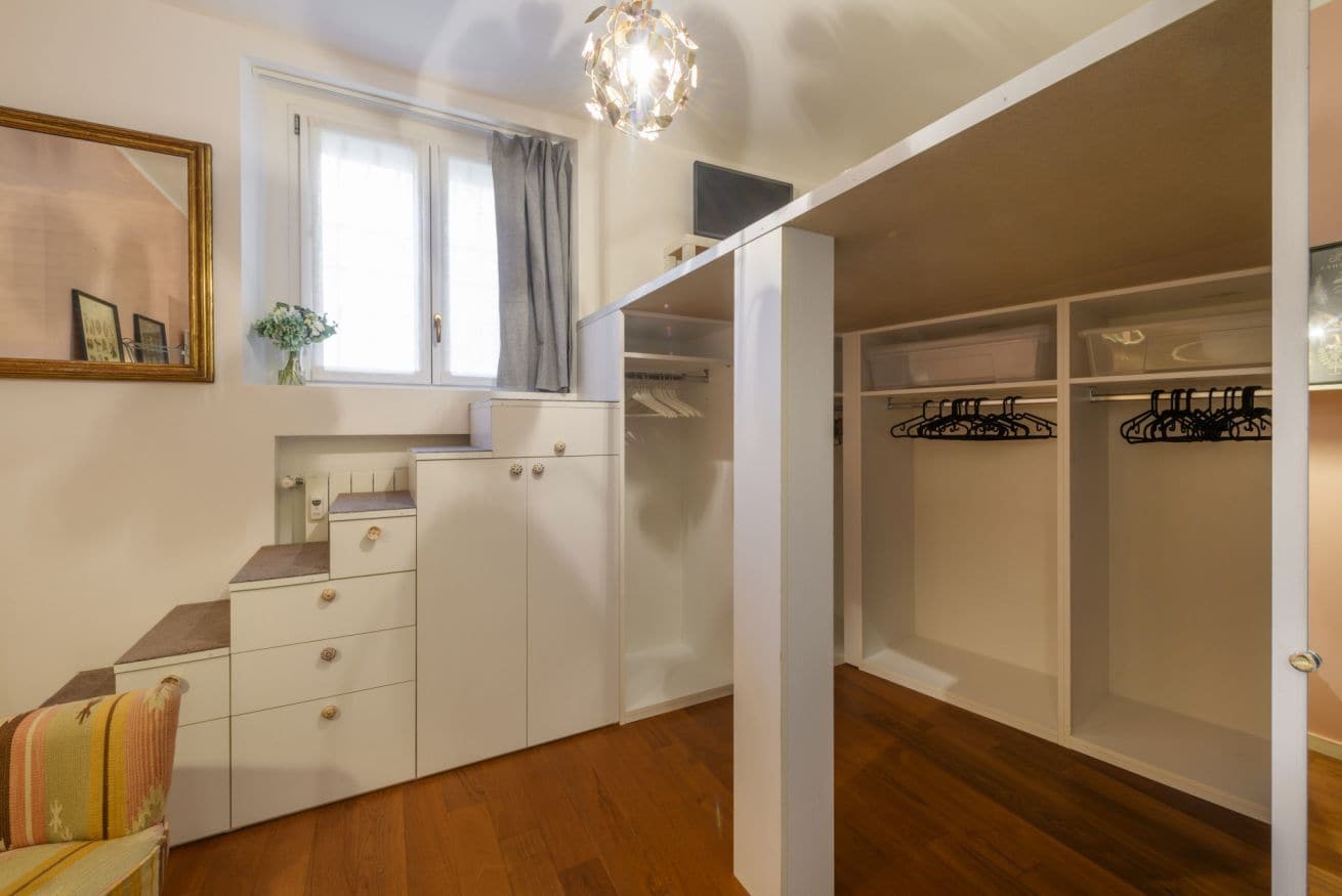 Una habitación con suelo de madera, un armario blanco, un escritorio pequeño, una silla, un ventanal con cortinas y una lámpara colgante.