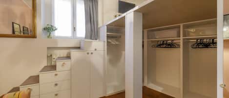 A room with a wooden floor, a white wardrobe, a small desk, a chair, a window with curtains, and a hanging light fixture.