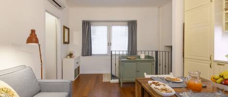 Une pièce comprenant un canapé, une table à manger avec des assiettes et un pichet, une fenêtre avec des rideaux et un buffet.