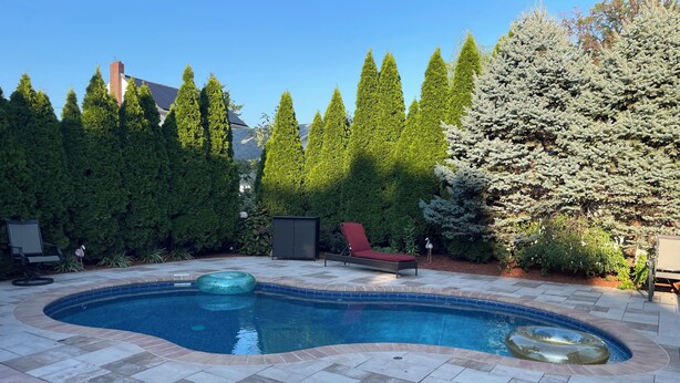 A swimming pool with a tiled deck, surrounded by evergreen trees and a red lounge chair. - 
Welcome to a comfortable, fully furnished 2-bedroom home located mins from  NYC (Bergenfield)