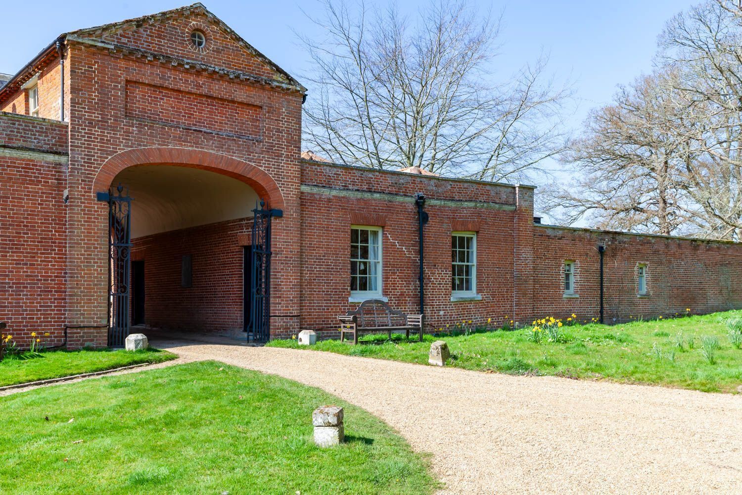 A brick building with a large arched entrance, a gravel path leading to it, and a bench outside.