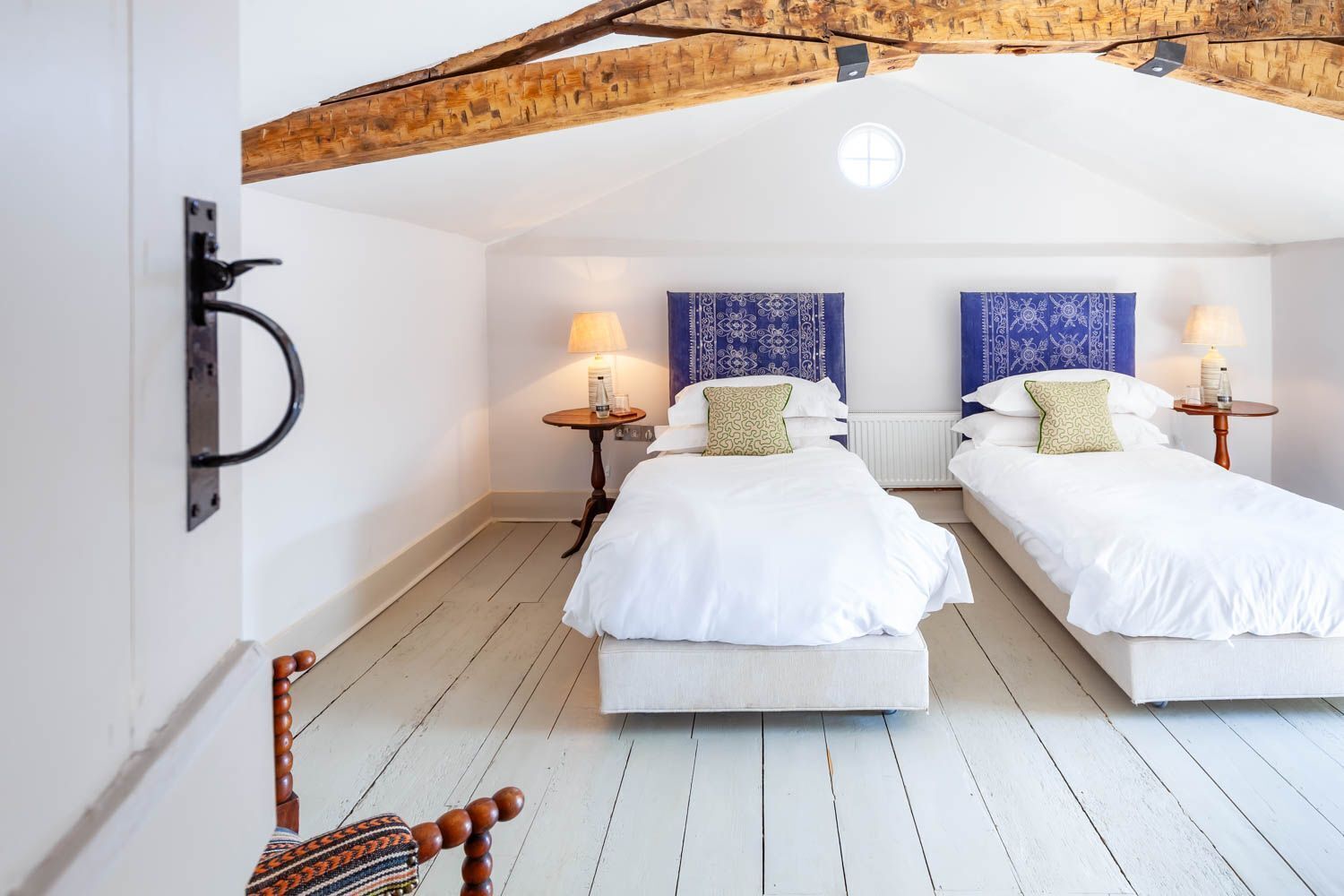Ein Zimmer mit zwei Betten, Holzbalken, einem kleinen Tisch mit Lampe und einem Stuhl.