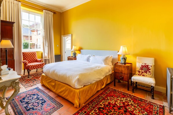 Une chambre d’hôtel avec un lit, deux chaises, une table de chevet, une lampe et une fenêtre avec des rideaux.