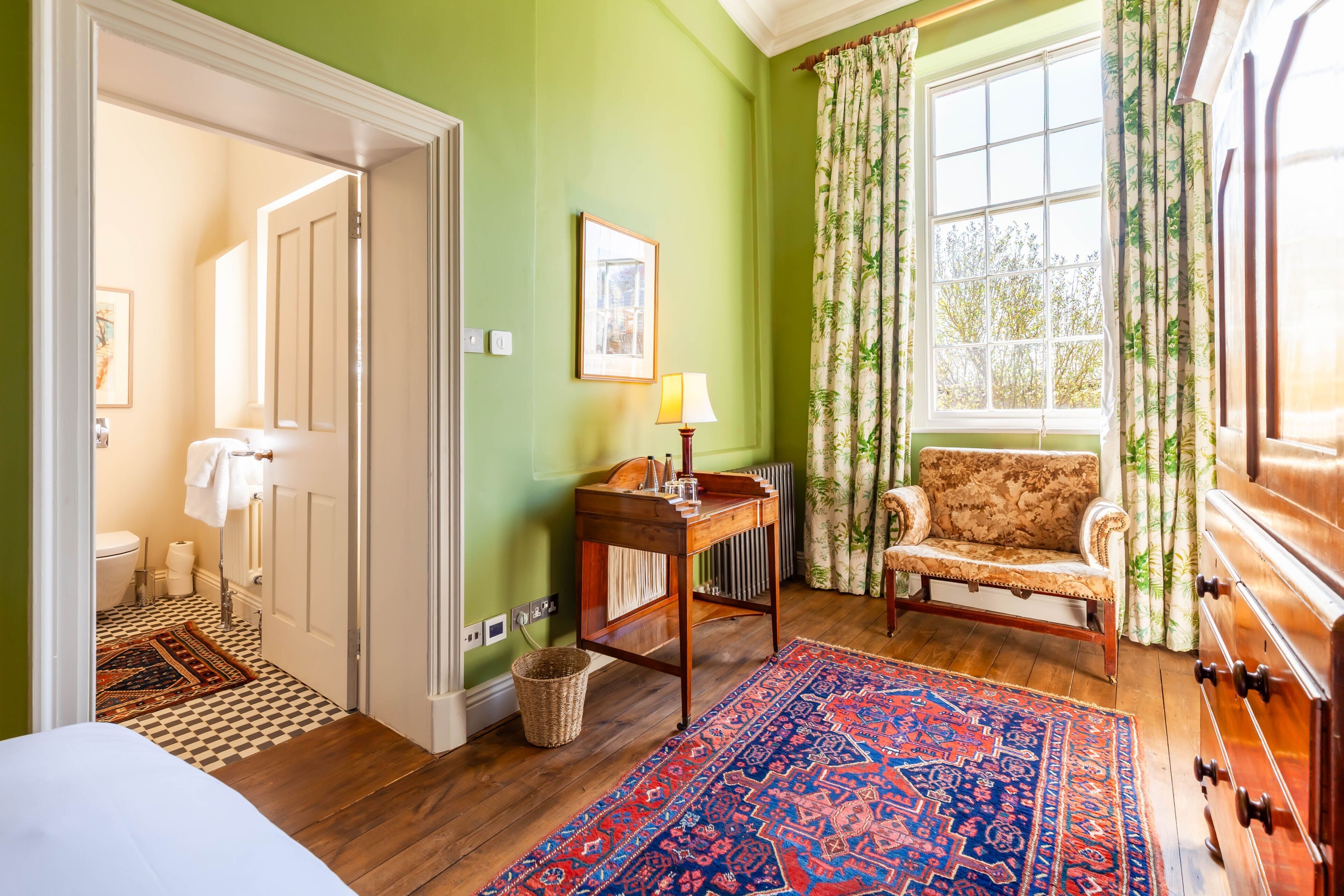 A room with a bed, a wooden dresser, a chair, a table with a lamp, a window with curtains, a radiator, and a patterned rug.