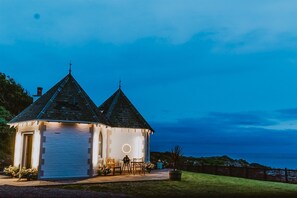 Un petit bâtiment blanc, doté d’un toit en pente, d’une fenêtre circulaire et d’une porte, situé dans un espace verdoyant et offrant une vue sur la mer.