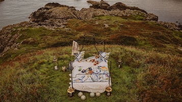 A bed with a floral bedspread, surrounded by grass and rocks, with a view of the water.