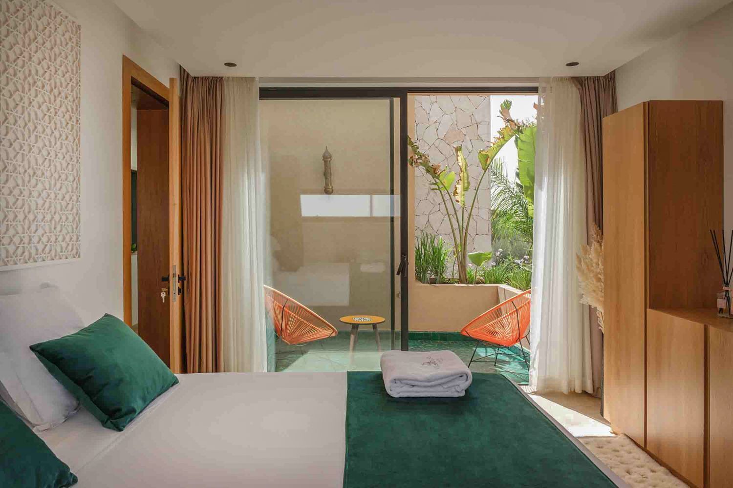 A hotel room with a bed, a green carpet, a small table, two orange chairs, and a view of greenery through a glass door.