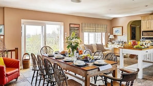 A dining area with a large table set for six, surrounded by chairs, with a view of the outdoors through a glass door. - Medlar House - Six Bedroom Villa, Sleeps 20 (Cheriton Fitzpaine)