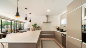 Een moderne keuken met een groot eiland, roestvrijstalen apparatuur, hanglampen en uitzicht naar buiten.