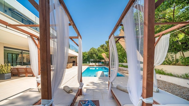 Een zwembadterras met een houten pergola, witte kussens en een tafel met een tijdschrift.