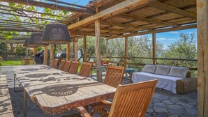 En udendørs terrasse med et langt træbord, stole, en sofa og et pergola med hængende lys.