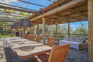 Patio esterno con un lungo tavolo in legno, sedie, un divano e una pergola con luci pendenti.